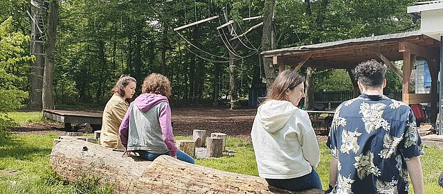 2023_Orgatag_ mehrere junge Menschen sitzen auf einem Baumstamm in der Natur sprechen